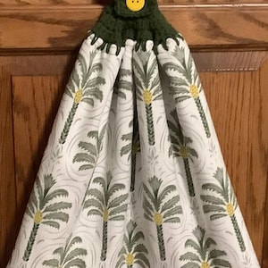 May include: A hanging kitchen towel with a green crocheted top and a yellow button. The towel features a white background with a repeating pattern of green palm trees and yellow accents. The towel is designed to hang on a cabinet handle.