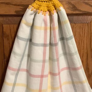 May include: A yellow crocheted dish towel holder with a green button. The holder is attached to a white dish towel with a plaid pattern in pink, yellow, and gray.