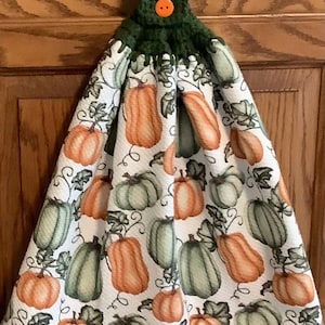 May include: A green crocheted dish towel with a button and a white background featuring a pattern of orange and green pumpkins.