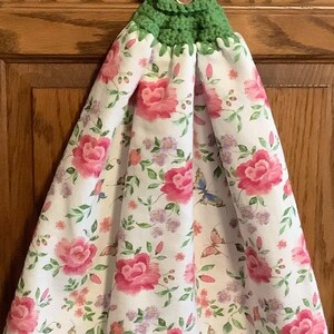 May include: A hanging kitchen towel with a white background, adorned with pink roses, purple flowers, and butterflies. The top is crocheted green with a button closure. A decorative and functional kitchen accessory.