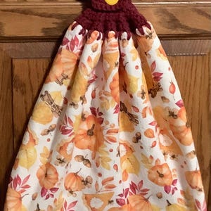 May include: A burgundy crocheted kitchen towel holder with a yellow button. The towel is white with a fall-themed print of orange pumpkins, red leaves, and cinnamon sticks.