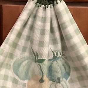 May include: A hanging kitchen towel with a green and white checkered pattern. The towel features a crochet top with a white button and a design of pumpkins in shades of blue and tan.