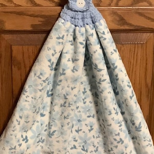 Puede incluir: Una lazada de colgar de ganchillo azul con un botón blanco está unida a una toalla de té blanca con un estampado floral azul.