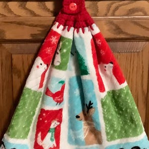 May include: A red crocheted kitchen towel topper with a button closure. The towel features a festive pattern with snowmen, reindeer, penguins, and a red bird on a white background with green and blue accents. A decorative kitchen accessory.