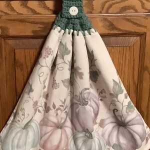 May include: A hanging kitchen towel with a green crocheted top and a white button. The towel features a pattern of pastel pumpkins in shades of blue, pink, and purple, with green leaves and vines on a cream background. A decorative fall-themed kitchen accessory.