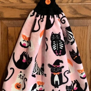 May include: A hanging kitchen towel featuring a light pink background with a repeating pattern of black cats, some wearing witch hats or orange collars, and white ghost figures holding pumpkins, secured by a black crocheted top with an orange button.