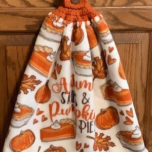 May include: A hanging kitchen towel featuring a white background with a repeating pattern of orange pumpkins, autumn leaves, and pumpkin pie slices. The text reads Autumn Skies & Pumpkin Pies. It has an orange crocheted top with a brown button.