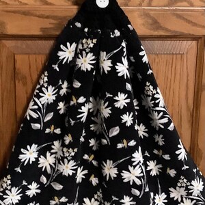 May include: A black and white daisy print kitchen towel with a black crocheted top and a white button. The towel features a repeating pattern of white daisies with yellow centers and green leaves.