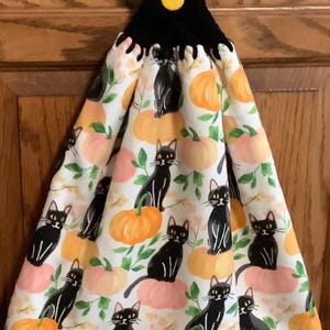 May include: A hanging kitchen towel featuring a white fabric with a repeating pattern of black cats and orange pumpkins. The top of the towel is gathered into a black crocheted topper secured with a single round yellow button for hanging.
