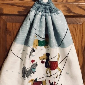 May include: A hanging kitchen towel with a light blue crocheted top and a white button. The towel features a winter scene with dogs wearing coats and hats, walking in the snow. The background is a light blue with white snowflakes.