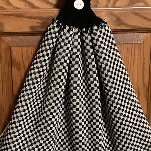 May include: A black and white checkered kitchen towel with a button closure. The towel has a black top with a white button and a checkered pattern. The bottom edge has a striped border. This decorative towel is designed to hang.