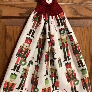 May include: A white kitchen towel with a red crocheted hanging loop and a white button. The towel features a repeating pattern of nutcracker soldiers with a dog's head.