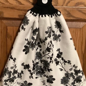 May include: A white kitchen towel with a black floral print and a black crocheted hanging loop with a white button.
