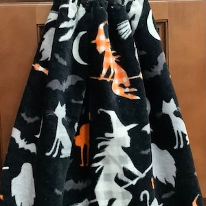 May include: Black kitchen towel with a Halloween theme. The towel features a white and orange witch, black cats, bats, and moons on a black background. The towel has a black crocheted loop for hanging.