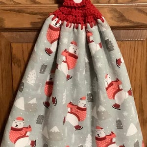 May include: A red crocheted kitchen towel topper with a white button. The towel features a light gray background with a pattern of polar bears wearing red sweaters and hats, ice skating among trees and mountains.