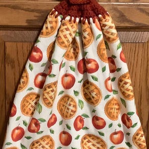 May include: A red crocheted kitchen towel holder with a yellow button. The towel is white with a pattern of apple pies and red apples.