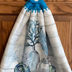 May include: A white kitchen towel with a blue crocheted hanging loop and a white button. The towel features a printed design of seashells, coral, and vases.