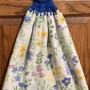 May include: A white kitchen towel with a floral pattern in shades of blue, yellow, and purple. The towel has a blue crocheted loop with a yellow button at the top.