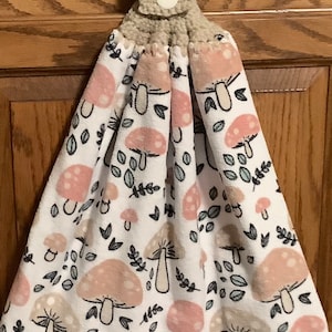 May include: A white kitchen towel with a mushroom and leaf pattern. The towel has a beige crocheted loop with a button for hanging.