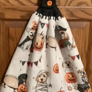 May include: A white hanging kitchen towel featuring a repeating pattern of dogs in various Halloween costumes, including a mummy, a pirate, and a taco, interspersed with orange pumpkins and black bunting, topped with a black crocheted hanger and orange button.