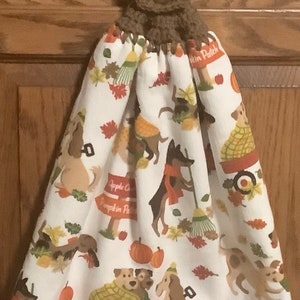 May include: A white kitchen towel with a fall-themed design featuring dogs, pumpkins, and leaves. The towel has a brown crocheted loop with a button at the top.
