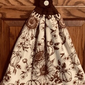 May include: A hanging kitchen towel featuring a white fabric base with a brown line-art print of sunflowers, pumpkins, and small flowers. The top section is made of dark brown crocheted yarn with a single white button for secure attachment.
