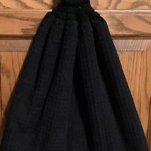 May include: A black kitchen towel with a textured waffle weave design. The top portion is crocheted with a button closure for hanging. The towel is hanging on a wooden door.