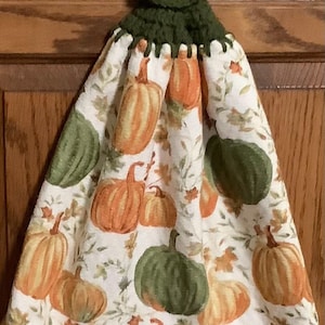 May include: A green crocheted dish towel holder with a yellow button. The towel is white with a pumpkin and leaf pattern.