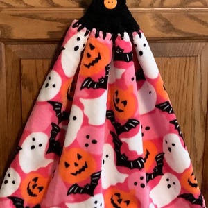May include: A Halloween-themed hanging kitchen towel. The towel features a pink background with white ghosts, orange jack-o'-lanterns, and black bats. The top is black crochet with an orange button.