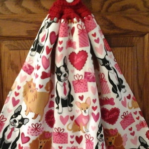 May include: A white tea towel with a red crocheted hanging loop and button. The towel features a Valentine's Day theme with pink hearts, presents, and dogs wearing bow ties. The dogs are a Boston Terrier, a Chihuahua, a Poodle, and a Shih Tzu.