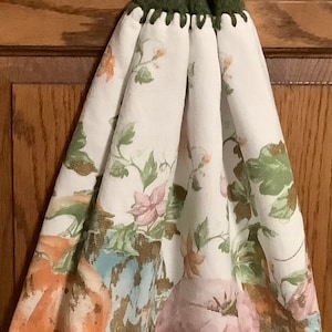 May include: A white kitchen towel with a floral pattern and a green crocheted hanging loop with a brown button. The towel features a design of pumpkins and other fall-themed elements.