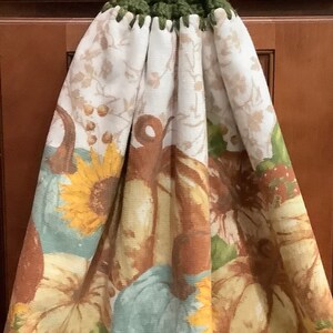 May include: A green crocheted dish towel with a brown button and a fall-themed print featuring pumpkins, sunflowers, and leaves.
