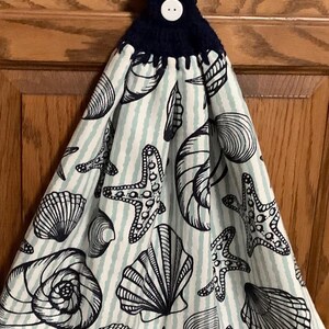 May include: A white kitchen towel with a blue and white striped pattern and a black and white seashell and starfish design. The towel has a navy blue crocheted loop with a white button.