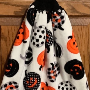 May include: A white kitchen towel with a black crocheted hanging loop and a button. The towel features a Halloween theme with orange, black, and white pumpkins.