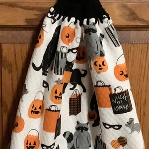 May include: A white kitchen towel with a Halloween theme. It features black cats, raccoons, pumpkins, and the words "trick or treat". The towel has a black crocheted loop with a button at the top.