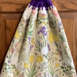 May include: A decorative kitchen towel with a purple crocheted top and a yellow button. The towel features a floral pattern with bunnies, ducks, and flowers in pastel colors, including yellow, pink, and purple.