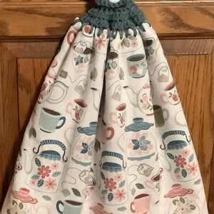 May include: A hanging kitchen towel with a white background and a repeating pattern of teapots, teacups, and tea bags in shades of pink, blue, and green. The top is a dark teal crochet with a white button.