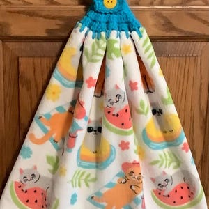 May include: A white hanging kitchen towel featuring a colorful pattern of cats lounging on watermelon slices and beach towels, accented with green leaves and small flowers. The towel is topped with a bright blue crocheted tab and a yellow button.
