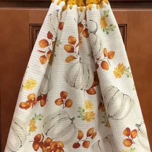 May include: A white kitchen towel with a pumpkin and leaf pattern. The towel has a mustard yellow crocheted loop with a brown button.