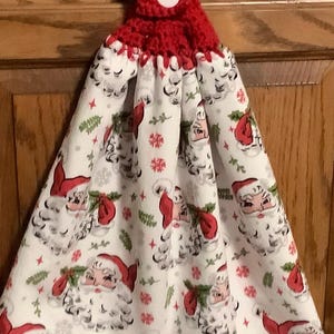 May include: A hanging kitchen towel featuring a white fabric with a repeating pattern of Santa Claus faces, red hats, and green holly leaves. The towel is secured at the top by a red crocheted topper with a single white button for hanging.