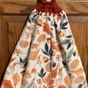 May include: A hanging kitchen towel featuring a white background with a repeating pattern of orange, brown, and dark blue autumn leaves and acorns. The top section consists of a rust-colored crocheted fabric with a single white button closure.