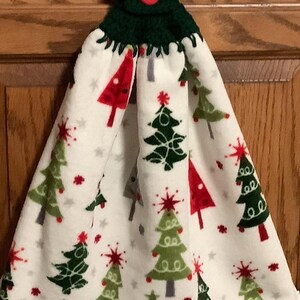 May include: A Christmas-themed hanging towel with a green crocheted top and a red button. The towel features a white background with repeating patterns of red, green, and gray Christmas trees and small stars. A festive kitchen accessory.