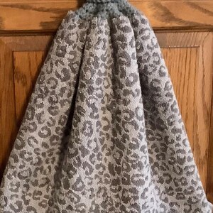 May include: A gray and white leopard print kitchen towel with a crocheted top and button closure. The towel is hanging on a wooden cabinet door. The towel is made of a soft, absorbent material.