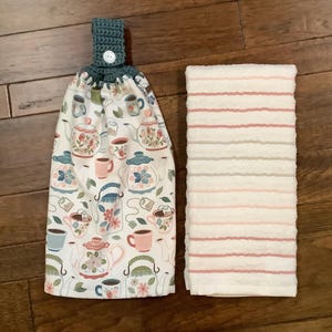 May include: A hanging kitchen towel with a teal crochet top and a button closure. The towel features a pattern of teapots, cups, and floral designs. A matching white and pink striped towel is next to it.