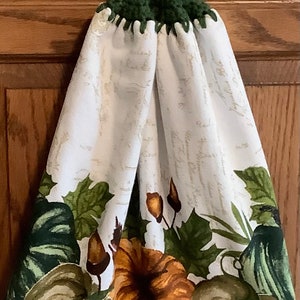 May include: A white kitchen towel with a green crocheted hanging loop and button. The towel features a fall-themed design with green and brown pumpkins and leaves.