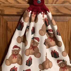 May include: A Christmas-themed kitchen towel with a white background featuring brown teddy bears wearing Santa hats and red bows. The towel also has green and red gift boxes. The top is a crocheted burgundy loop with a button.