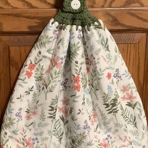 May include: A hanging kitchen towel with a white floral pattern featuring pink, red, and purple flowers and green leaves. The top is crocheted in green with a white button for hanging. The towel is designed to hang on a cabinet handle.