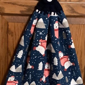 May include: A navy blue hanging kitchen towel with a black crocheted top and white button. The towel features a repeating pattern of polar bears wearing red scarves and hats, along with white snowflakes, gray mountains, and green trees.