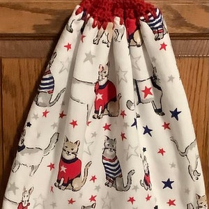 May include: A white kitchen towel with a red, white, and blue cat pattern. The towel has a red crocheted loop with a white button at the top.