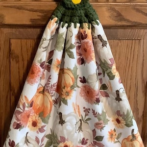 May include: A green crocheted dish towel hanger with a yellow button. The towel is white with a floral pattern featuring pumpkins, leaves, and flowers in shades of orange, brown, and yellow.